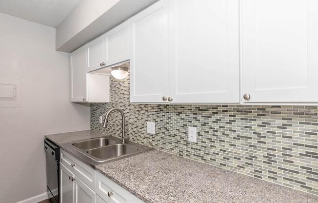 Modern kitchen featuring white cabinets, a stainless steel sink with a gooseneck faucet, and a sleek countertop. The backsplash consists of textured tiles in shades of gray and green, complemented by under-cabinet lighting. A black dishwasher is also visible in the corner.