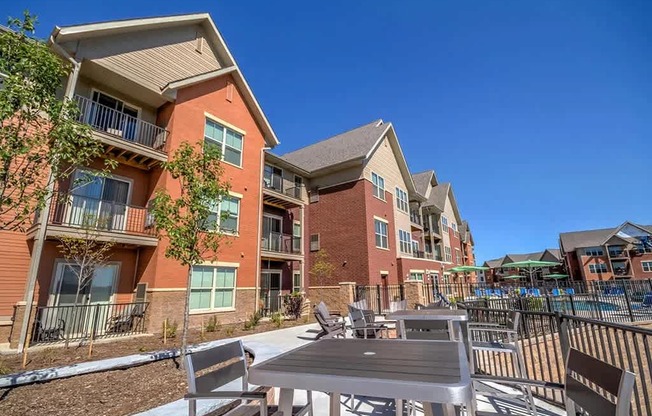 A sunny outdoor patio here at Vue at Pinnacle Park with dining tables and chairs, nearby apartment buildings with balconies, landscaped walkways, and a pool area in view for relaxed community gathering.