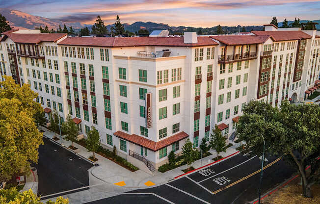 Corner view of a modern apartment exterior with tile rooflines, warm architecture, and tree-lined surroundings