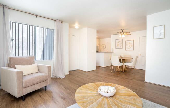 A spacious living area featuring light wood flooring, a beige armchair with a cushion, and a round table with chairs. Large windows with curtains allow natural light, and a clock and framed artwork decorate the walls. The kitchen area is visible in the background.