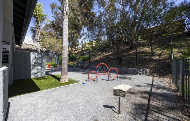 A dog park area featuring three red agility hoops on gravel, bordered by a fence and surrounded by greenery. There is a sign that reads "People please clean up after your pet." The park is sunny, with palm and other trees lining the slope behind the fenced area.