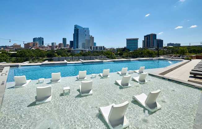 A pool with white chairs and a city skyline in the background.