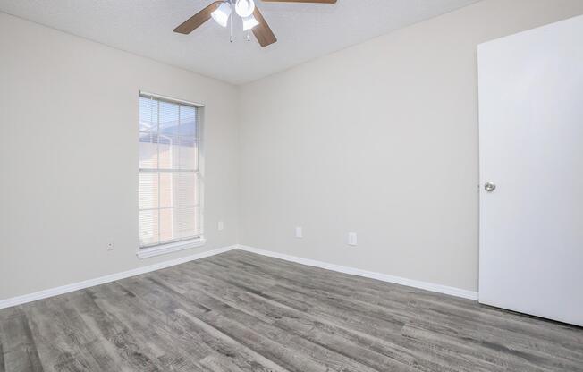 A light-colored room featuring a ceiling fan, large window with blinds, and hardwood-style flooring. The walls are painted in a neutral tone, and there is a closed white door on the right. The room is empty, providing a spacious feel.