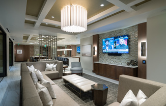 A seating area adjacent to the clubhouse kitchen and dining area with a large wraparound sectional couch in front of an HDTV.