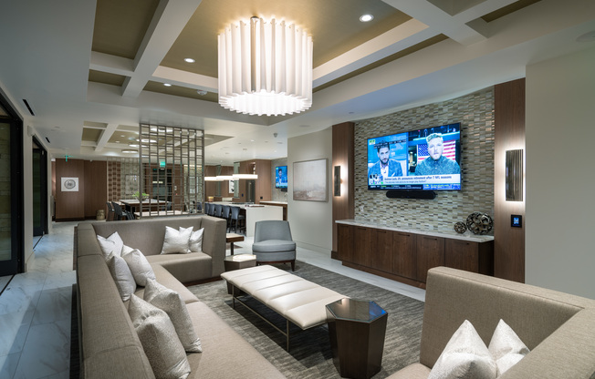 A seating area adjacent to the clubhouse kitchen and dining area with a large wraparound sectional couch in front of an HDTV.