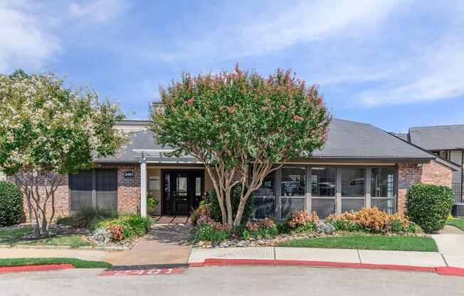A modern brick building with large windows and a welcoming entrance, surrounded by well-maintained landscaping featuring flowering bushes and trees, under a blue sky with scattered clouds. Pathway leading to the entrance is lined with colorful flowers and greenery.