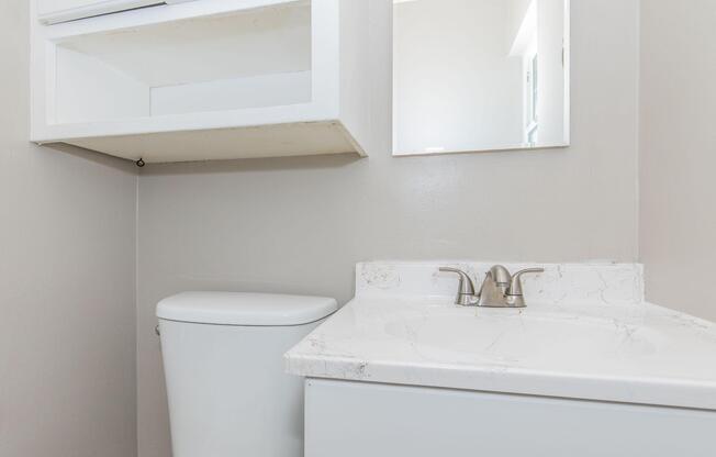 A clean and simple bathroom featuring a white toilet, a sink with a marble countertop, and a wall mirror. The walls are painted in a light color, and there is a shelf above the sink for storage. The overall design is minimalistic and functional.