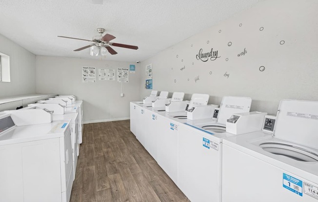 A room with a white ceiling and floor with a wall of washing machines on the right and a fan on the ceiling.