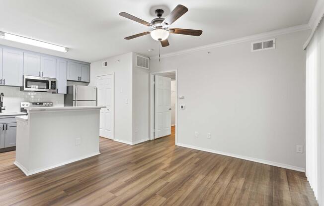 Modern kitchen and living area featuring wood flooring, a ceiling fan, and a light color palette. The kitchen includes gray cabinets, stainless steel appliances, and a countertop that divides the space. A door leads to another room, while large windows provide natural light.