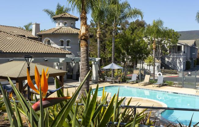 A peaceful residential area featuring a swimming pool surrounded by lush greenery, palm trees, and tropical plants. In the background, a charming building with a tiled roof and an outdoor gazebo creates an inviting atmosphere for relaxation and leisure.