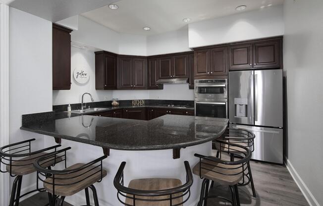 Modern kitchen featuring dark wood cabinets, a black granite countertop, and stainless steel appliances. The space includes a large island with bar stools and a decorative sign on the wall. Bright lighting enhances the clean and inviting atmosphere of the kitchen.