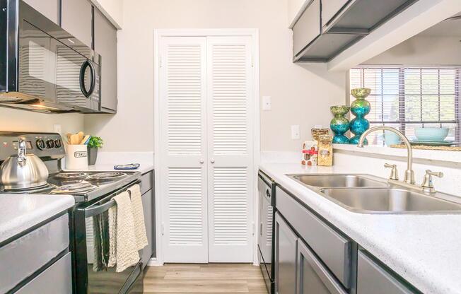 Bright kitchen featuring dark cabinets, stainless steel appliances, and a white countertop. Includes a double sink with a modern faucet, a decorative glass bowl, and a pantry door with louvered panels. Sunlight streams through a window, enhancing the inviting atmosphere.