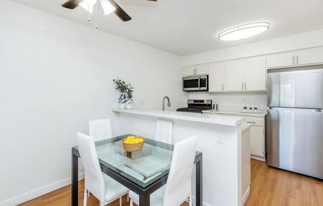 A modern kitchen featuring a dining area with a glass table and white chairs. The kitchen has sleek white cabinets, stainless steel appliances, and a bowl of lemons as a centerpiece. Natural light illuminates the space from a ceiling fan and overhead light fixture. The floor is wooden, enhancing the contemporary feel.