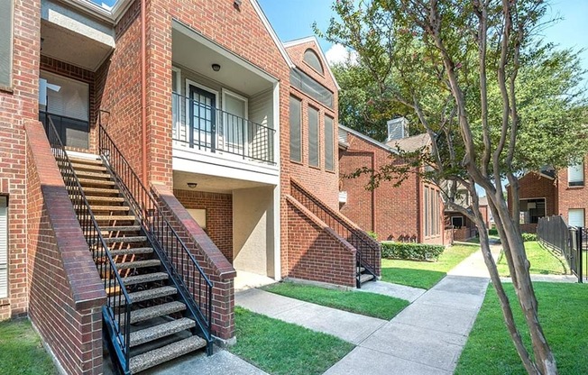 A landscaped apartment exterior here at Lakewood on the Trail with brick buildings, private balconies, outdoor staircases, and green walkways, creating a clean and welcoming residential setting.