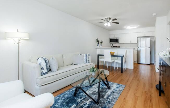A bright and modern living area featuring a light-colored sofa with blue and white decorative pillows, a glass coffee table on a blue area rug, a dining table with four chairs, and a kitchen area with stainless steel appliances. The walls are white, and there is a ceiling fan and warm wooden flooring.