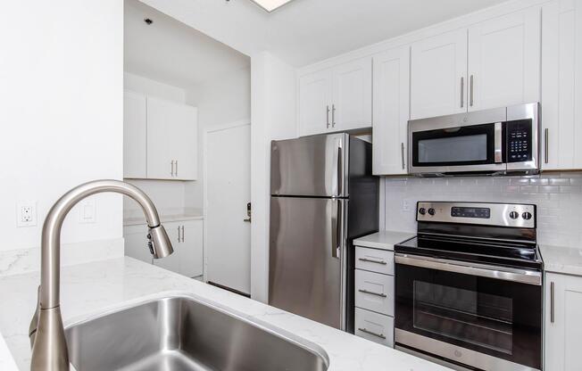 Modern kitchen featuring stainless steel appliances, including a refrigerator, microwave, and oven. The cabinets are white with sleek handles, and the countertop is made of light-colored stone. A stainless steel sink is visible in the foreground, with a view towards a door leading to another area.