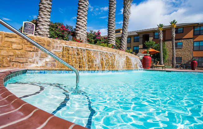 A refreshing resort-style swimming pool here at Tuscany at Faudree featuring clear blue water, a shallow entry with handrail, and a stone waterfall feature. Tall palm trees, colorful landscaping, and Mediterranean-inspired apartment buildings surround the pool beneath a bright blue sky.