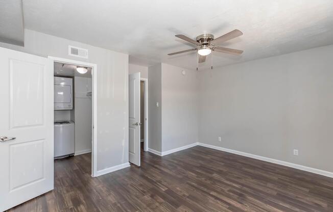 A spacious, empty room with light gray walls and dark wooden flooring. A ceiling fan is installed, and there are two doors: one leading to a laundry area and another to an adjoining room. The natural light fills the space, making it bright and inviting.