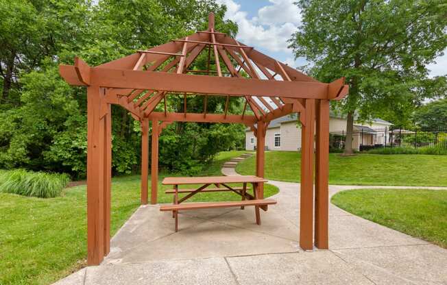Wood gazebo with picnic table next to walkway surrounded by green grass and mature trees