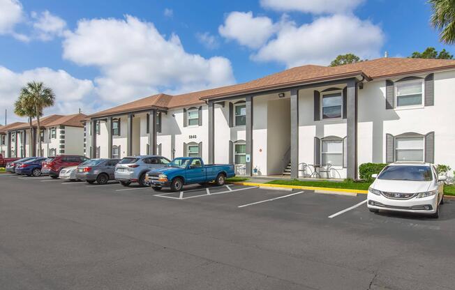 A white two-story apartment building with brown shingles, featuring multiple parking spaces filled with various cars. A clear blue sky with scattered clouds is overhead. The scene includes palm trees and well-maintained landscaping.
