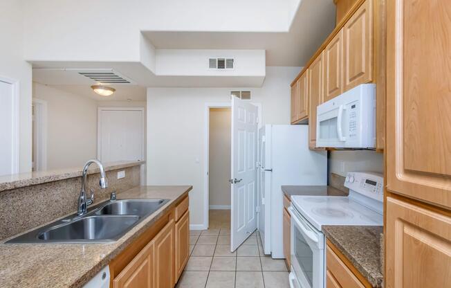 A modern kitchen featuring light wooden cabinetry, a double sink, and a countertop. Appliances include a microwave, an oven, and a refrigerator. The room has tiled flooring and a door leading to another room. Natural light brightens the space, creating a welcoming atmosphere.