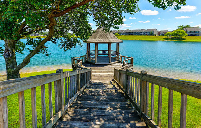 A wooden walkway leads to a gazebo by a lake at Somerset Lakes Apartments, offering a tranquil outdoor retreat.