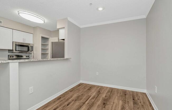 A modern kitchen and dining area in a well-lit apartment. The space features light gray walls, wooden flooring, and a bar counter with a countertop. A microwave and kitchen cabinets are visible, creating a cozy and inviting atmosphere. Bright overhead lighting enhances the open layout.