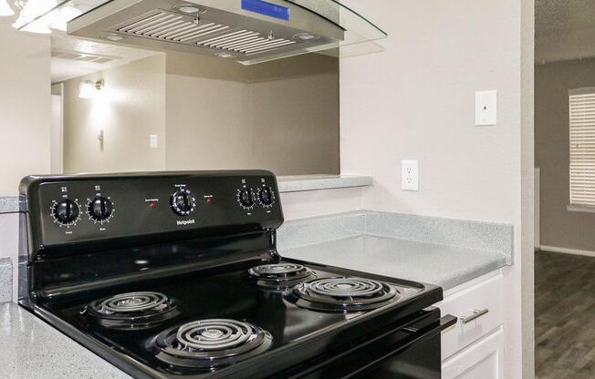 A close-up view of a black electric stovetop with four burners, situated on a gray textured countertop. A kitchen hood is installed above the stove. The background shows a light-colored wall and a window with blinds, indicating a well-lit, modern kitchen area.