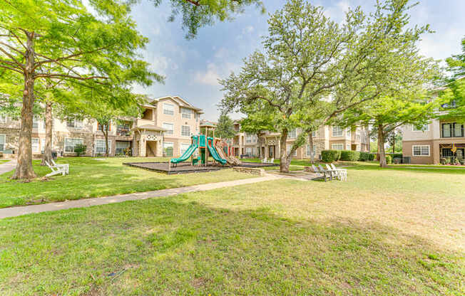 A playground with a green slide is surrounded by trees and grass in a residential area.