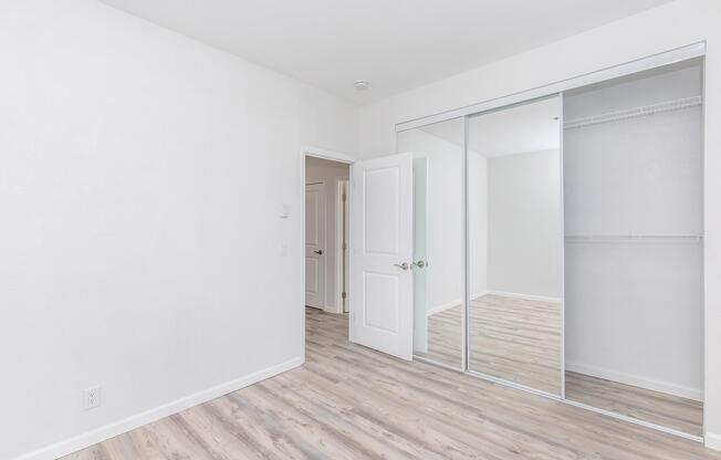 A light-colored room with white walls and hardwood-style flooring. It features a large mirrored closet with sliding doors, an open doorway leading to another room, and no furniture. The space appears bright and minimalistic, suitable for various uses.