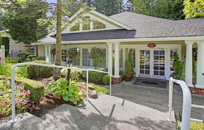 A welcoming leasing office exterior here at Aspen Creek featuring a charming single-story building with white columns, large windows, and double glass entry doors, surrounded by manicured landscaping, flowering plants, hanging baskets, and a gently sloped walkway with handrails leading to the main entrance.