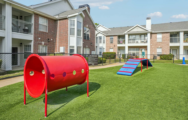A red tunnel-like structure sits in the middle of a grassy area in front of apartment buildings.