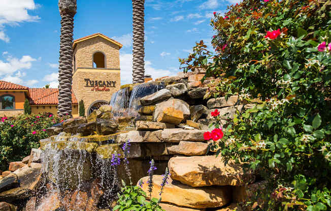 A scenic stone waterfall feature here at Tuscany at Faudree framed by lush greenery, blooming red roses, and textured rock tiers. Flowing water cascades over natural stone beneath tall palm trees, with Mediterranean-style buildings and a blue sky backdrop creating a serene, resort-inspired community entrance.