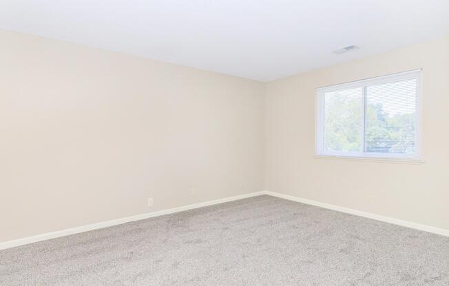 Empty room with light beige walls and a carpeted floor. A single window on the left side allows natural light to enter, illuminating the space. The room appears spacious and uncluttered, creating a neutral canvas for personal decoration.