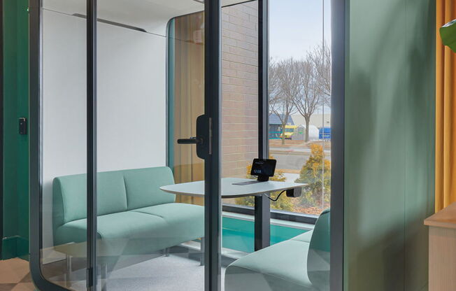 A glass door separates a room with a green chair and table from another room.
