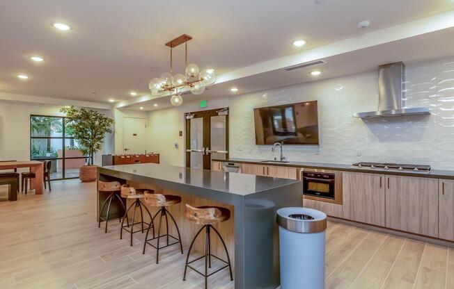 Modern kitchen and dining area featuring a sleek island with seating, stylish light fixtures, a large window with natural light, and contemporary appliances. The space is designed with wood and neutral tones, complemented by a potted plant and a trash bin, creating a warm and inviting atmosphere.