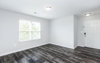 A bright, empty room with light gray walls and dark wooden flooring. There is a window on the left side, allowing natural light in, and a white door on the right leading outside. The space is well-lit with overhead fixtures, creating a clean and modern atmosphere.