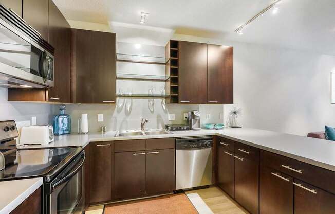 A modern kitchen here at Blue and Lime featuring rich dark wood cabinetry, light countertops, and a clean, functional layout. Stainless steel appliances, a double sink, open glass shelving, and track lighting add visual interest, while the spacious counters and thoughtful storage create an inviting space for everyday cooking and entertaining.