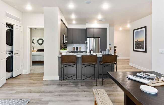 A contemporary open kitchen here at Reflect at Dobson Ranch featuring dark gray cabinetry, a granite-style breakfast bar with seating, stainless-steel appliances, and wood-style flooring, with recessed lighting overhead and an open layout that connects seamlessly to the dining area and nearby living space.
