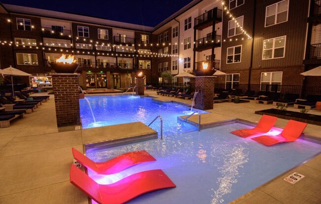 the pool is lit up at night with red chairs