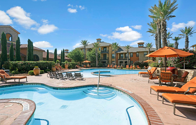 A bright resort-style swimming pool here at Tuscany at Faudree featuring a shallow entry area, curved pool design, and surrounding tan pool deck with orange lounge chairs and umbrellas. Palm trees, landscaped greenery, and Mediterranean-inspired apartment buildings frame the space under a clear blue sky.