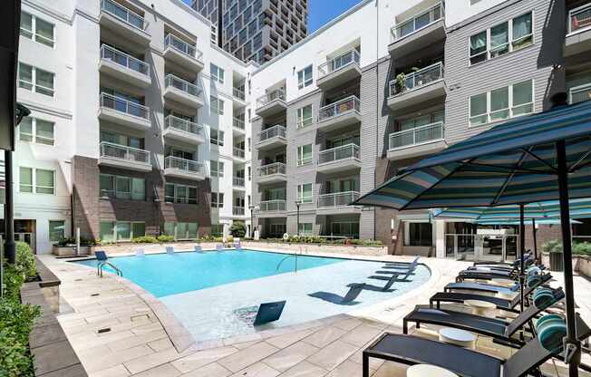 A swimming pool surrounded by lounge chairs and umbrellas in front of apartment buildings.