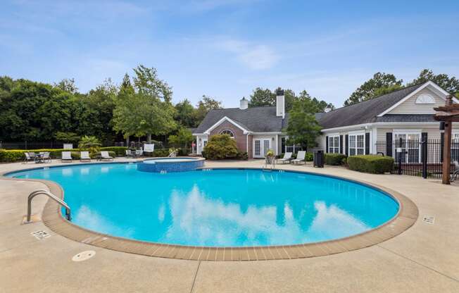 Luxury apartment community outdoor pool with crystal-clear water, surrounded by a spacious sundeck with lounge chairs. A brick and white clubhouse with large windows and landscaping is in the background, along with lush green trees under a bright blue sky