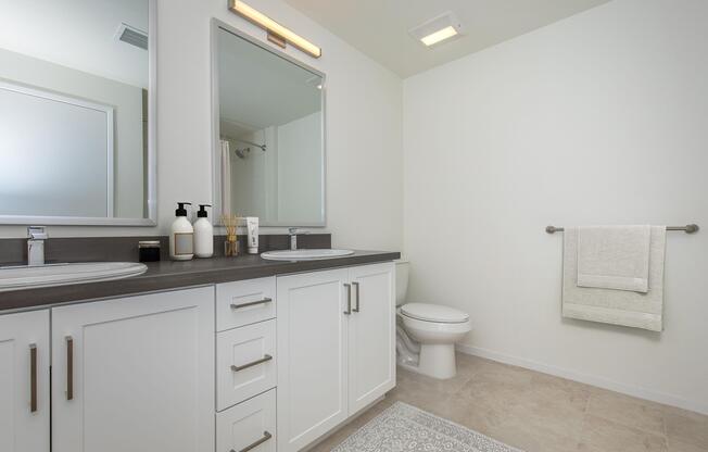 A modern bathroom featuring dual sinks with mirrors, a dark countertop, a white vanity with storage, a contemporary toilet, and a neatly hung towel. Soft lighting illuminates the space, which has neutral-colored walls and tiled flooring. A rug adds a touch of warmth to the room.