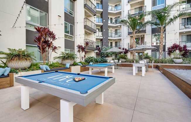 A pool table with a blue cloth and white legs is in the middle of a courtyard with plants and a building in the background.
