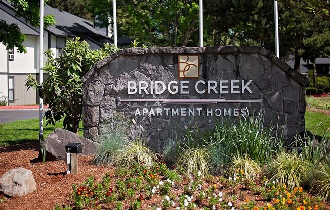 A sign for Bridge Creek Apartment Homes is surrounded by flowers and plants.