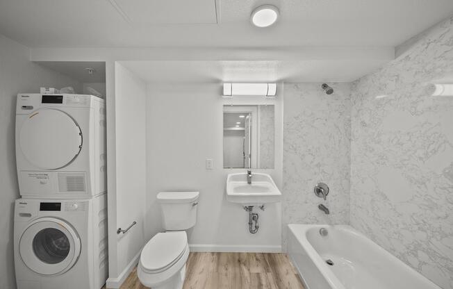 A modern bathroom featuring a white bathtub, a sink with a wall-mounted faucet, and a toilet. To the left, there is a stacked washing machine and dryer. The space has light marble walls, a large mirror, and wooden flooring, creating a clean and contemporary aesthetic.