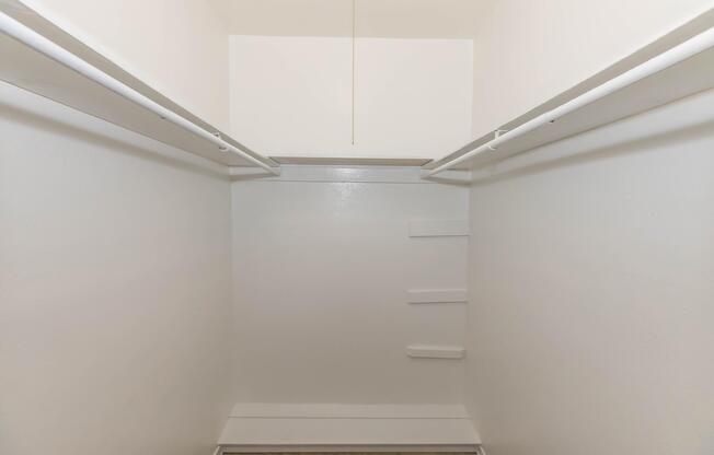Empty closet with two hanging rods along the sides and a single light fixture above. The walls are painted white, and there are a few shelves on one side. The floor is carpeted, giving a clean and minimalistic appearance.