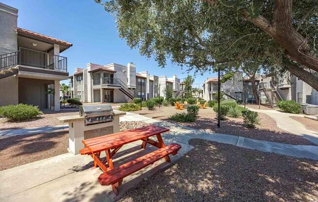 A red picnic table is in the middle of a gravel area with houses in the background.