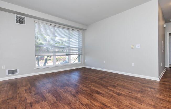 A spacious, empty living room featuring a large window with blinds, light gray walls, and brown laminate flooring. The room has minimal furnishings and a serene atmosphere, creating a bright and open space ideal for personalization.